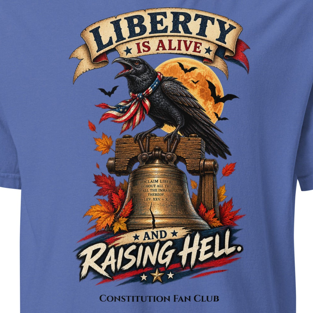 A close-up of a blue graphic t-shirt featuring a raven perched on a Liberty Bell with autumn leaves, a full moon, and banners reading 'LIBERTY IS ALIVE' and 'AND RAISING HELL.' The text 'CONSTITUTION FAN CLUB' is printed below.